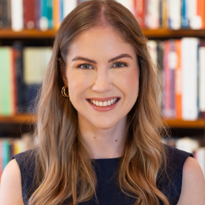 Fotografia de Arielle Sagrillo em fundo com estante de livros desfocada: mulher adulta sorrindo, pele clara, cabelo comprido castanho-claro ondulado. Usa brincos dourados pequenos. Veste blusa preta sem mangas. Expressão alegre e amigável.