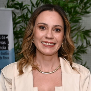 Fotografia de Marcela Novais em ambiente interno: mulher adulta sorrindo, pele clara, cabelo ondulado castanho com mechas mais claras até os ombros. Usa brincos pequenos e colar metálico. Veste blazer bege claro; ao fundo há plantas e um banner com texto pouco legível.