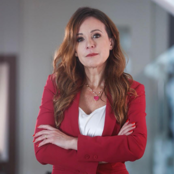 Fotografia de Valeria Scarance em ambiente interno: mulher adulta, pele clara, cabelo longo ruivo ondulado. Está de braços cruzados, com expressão neutra e batom vermelho. Usa blazer vermelho sobre blusa branca e colar com pingente rosa. Fundo desfocado.