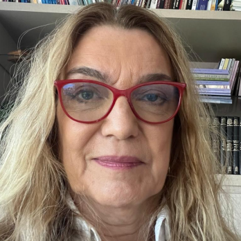 Fotografia de Vivian Pares em ambiente interno com estante de livros ao fundo: mulher adulta, pele clara, expressão neutra com leve sorriso, cabelo loiro-escuro com mechas mais claras, solto e levemente ondulado, com risca próxima ao centro. Usa óculos grandes de armação vermelha e batom em tom rosado. Veste camisa branca.